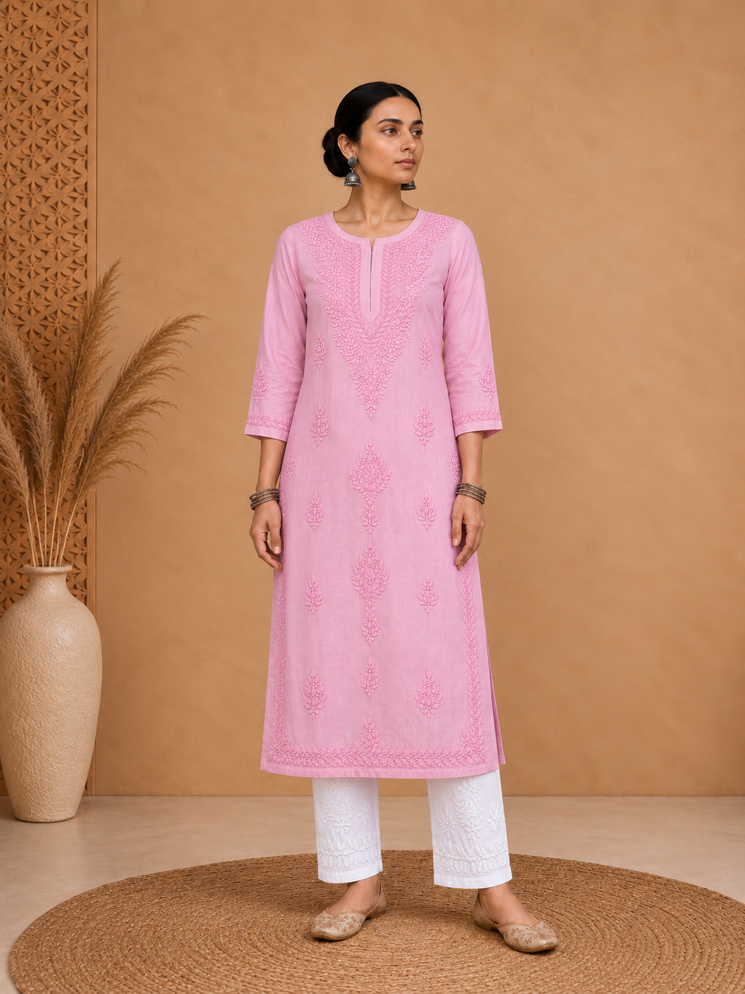 A woman stands indoors wearing a light pink embroidered kurta with white pants and beige flat shoes, posed against a warm tan wall with a woven rug and a vase with dried grass.